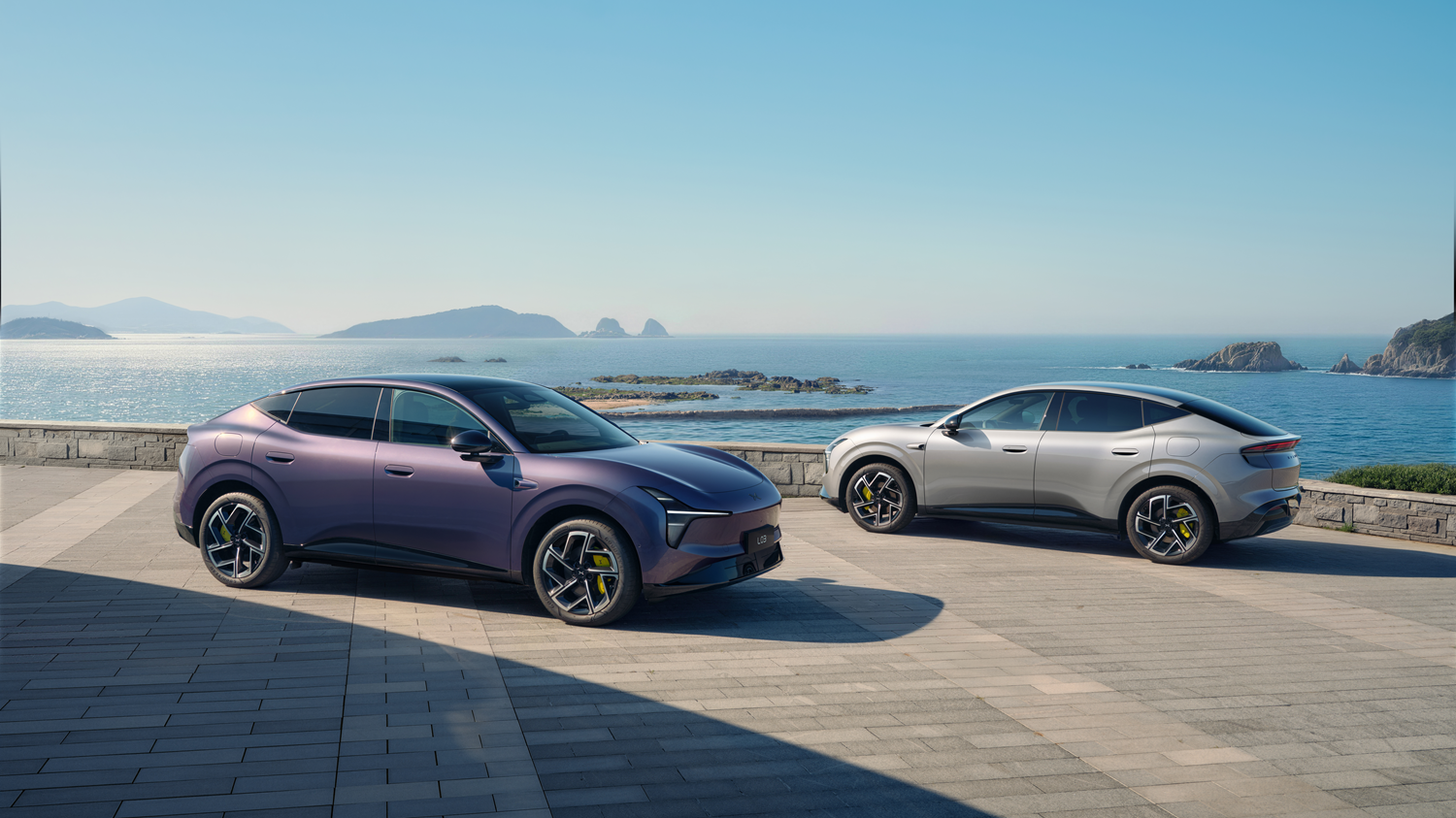 Zwei XPENG L03 Elektrofahrzeuge in violetter sowie grauer Lackierung an der Küste.